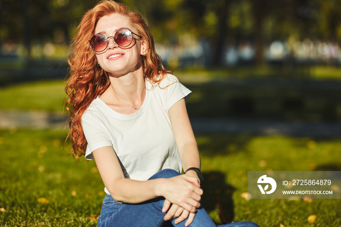 A lovely red-haired girl, warmed by the rays of the sun, is sitting on a lawn and posing for the cam