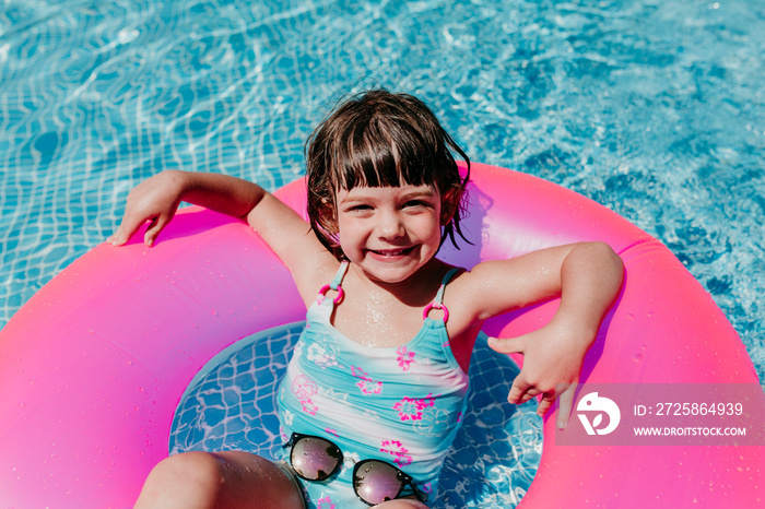 美丽的小女孩漂浮在游泳池里的粉色甜甜圈上。戴着墨镜，面带微笑。有趣又夏天