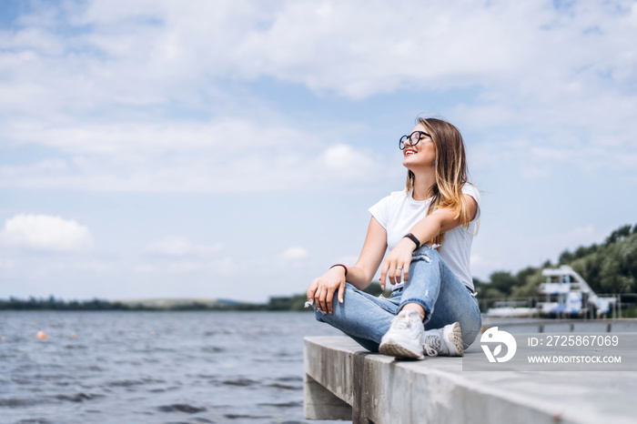 Young woman with long hair in stylish glasses posing on the concrete shore near the lake. Girl dress