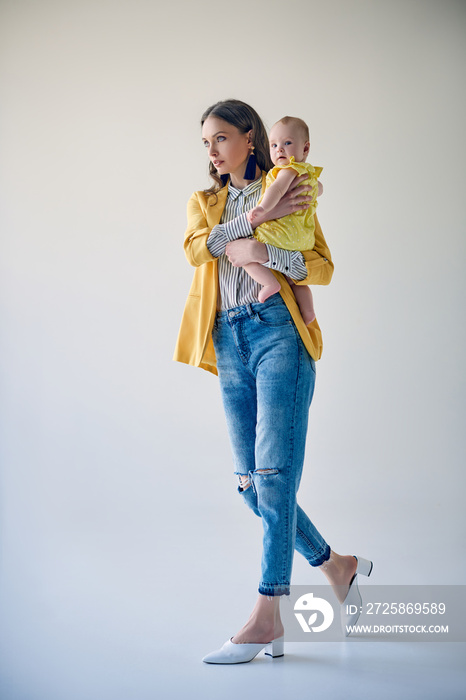 full length view of stylish young mother carrying adorable baby daughter and walking on grey