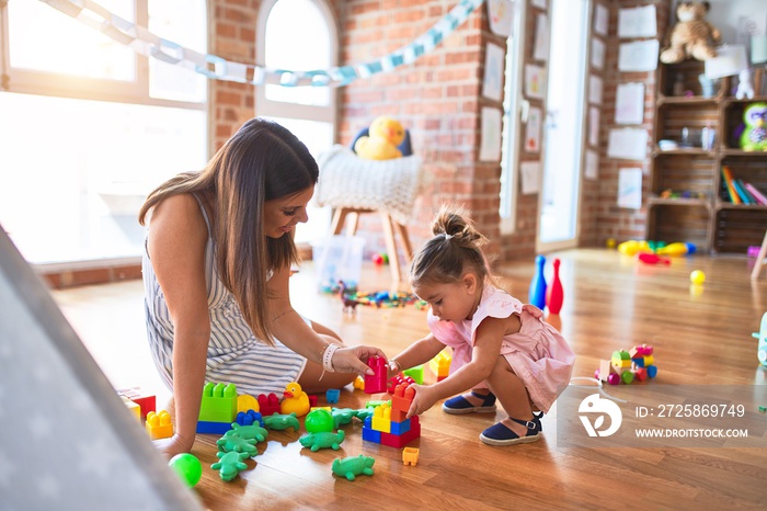 Young beautiful teacher and toddler sitting on the floor playing with building blocks toy at kinderg