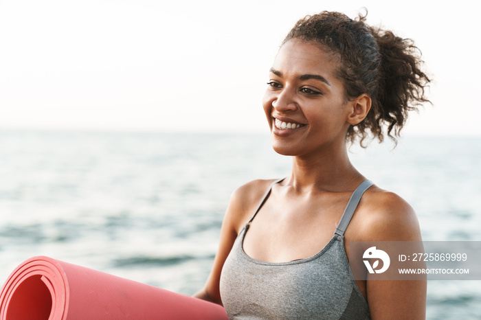 Photo of joyful african american sportswoman smiling and standing with yoga mat by seaside in mornin