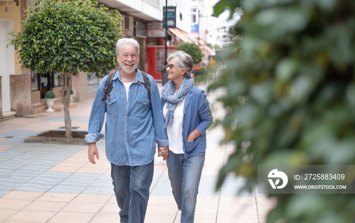 Happy active smiling adult Caucasian senior couple walking in the city holding hands as tourists. At