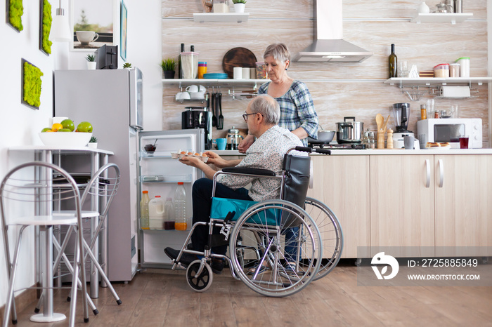 Disabled senior male in wheelchair taking eggs carton from refrigerator helping his wife in kitchen.