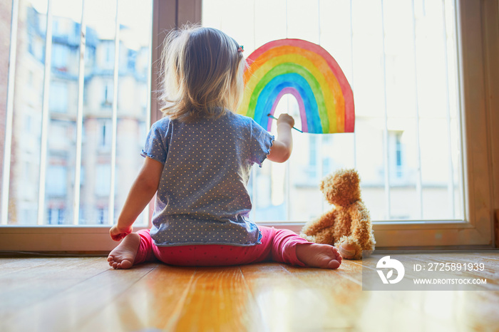 Adorable toddler girl painting rainbow on the window glass