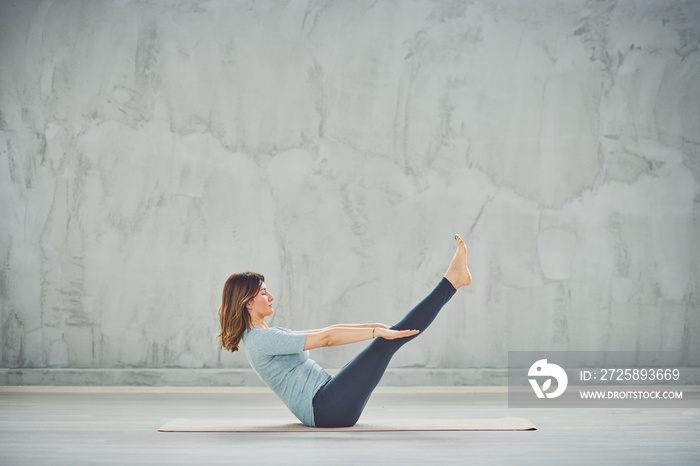 Beautiful Caucasian brunette in blue sportswear sitting on the mat in boat yoga position.