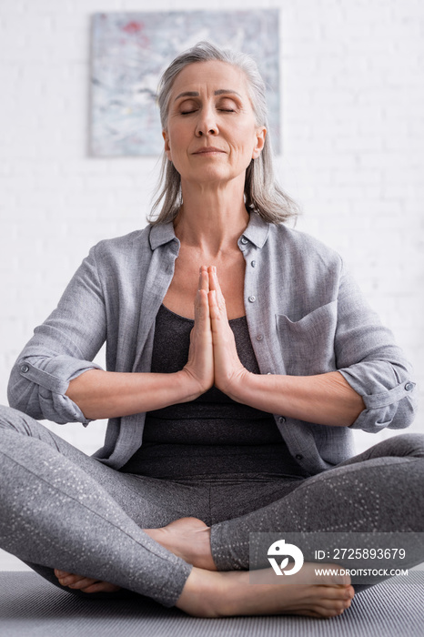 mature woman with praying hands and closed eyes sitting in lotus pose on yoga mat