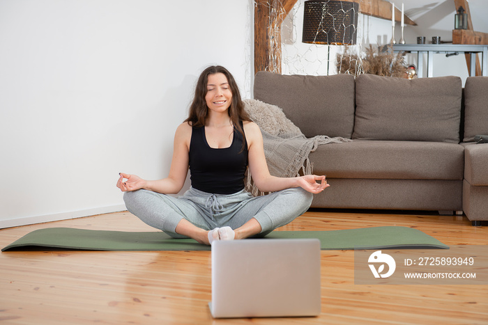 瑜伽在线。放松的年轻女子在家里的笔记本电脑前冥想，以莲花姿势坐着
