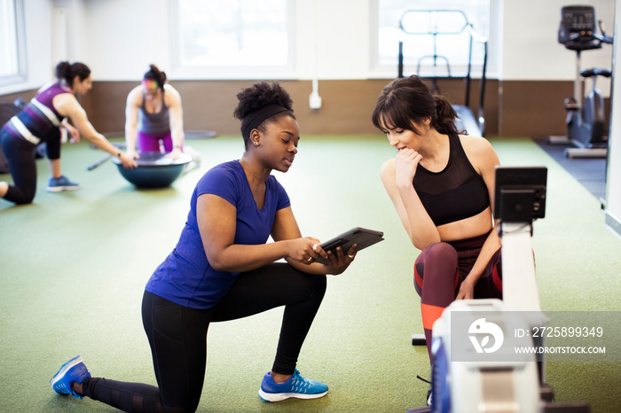 Instructor showing tablet computer to female athlete exercising on rowing machine in gym