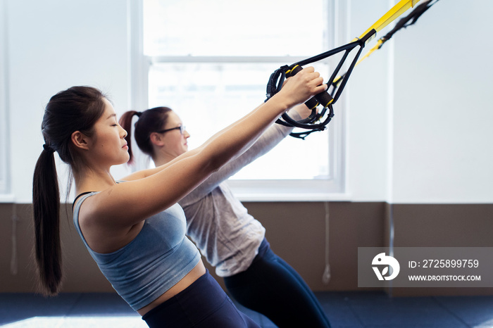 Side view of confident female athletes pulling suspension straps against windows in gym