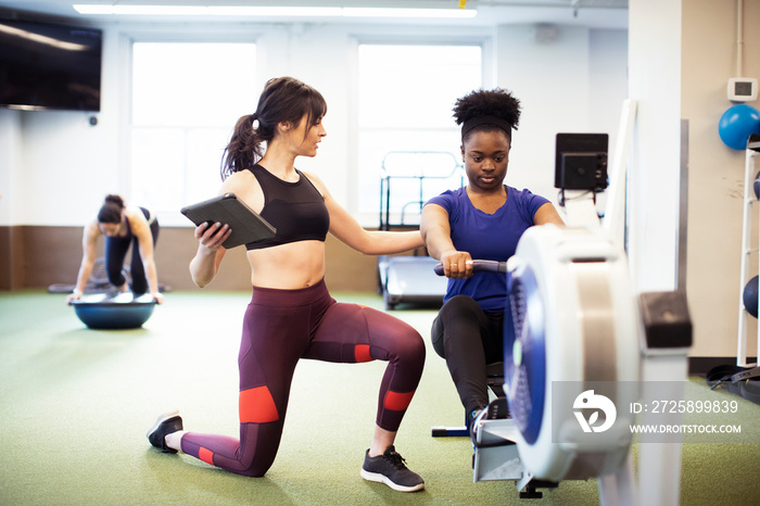 Instructor with tablet computer guiding athlete exercising on rowing machine in gym