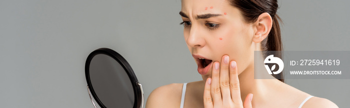 panoramic shot of shocked woman with acne on face looking at mirror isolated on grey