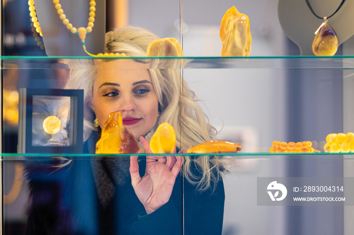 A young, beautiful, blonde woman looks at amber jewelry at a jewelry store. View through the window 