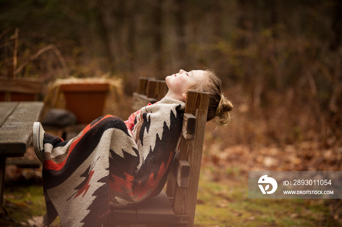 Side view of girl with eyes closed wrapped in blanket relaxing on bench at forest