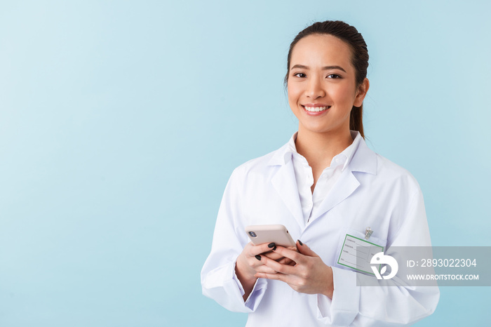 Woman doctor posing isolated over blue wall background using mobile phone.