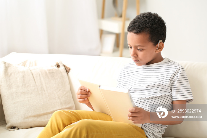 Little African-American boy with hearing aid reading book at home