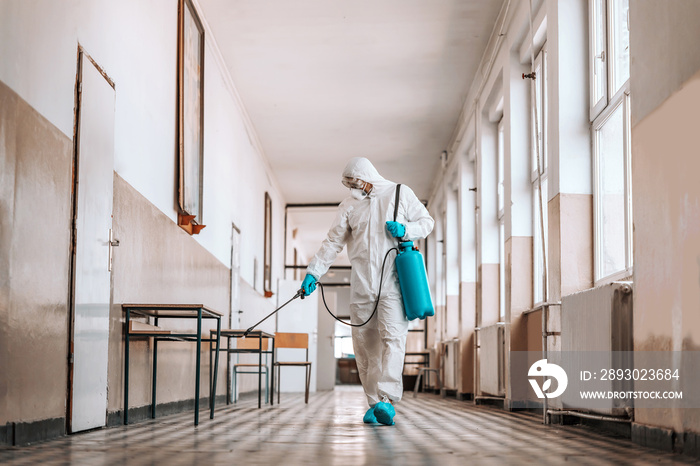 Worker in sterile white uniform, with mask and glasses holding sprayer with disinfectant and sprayin