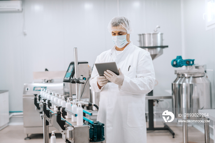 Portrait of Caucasian chemist in sterile uniform using tablet while standing next to machine with li