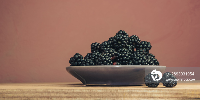 Fresh blackberry in bowl on a brown wooden table and dark-red wall background.