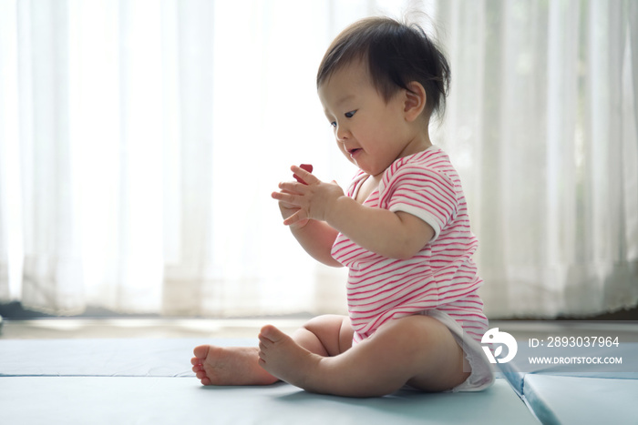 Asian cute baby sitting and playing a small toy on soft mat at home. The kid holding the toy and que