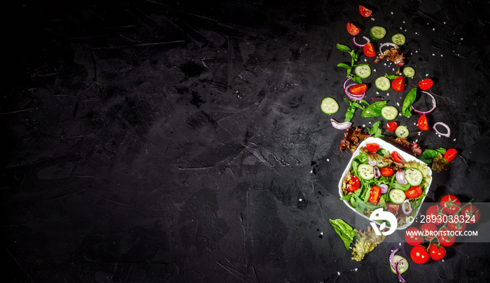 Various fresh mix salad leaves with tomato and cucumber in a glass bowl on dark background with ingr