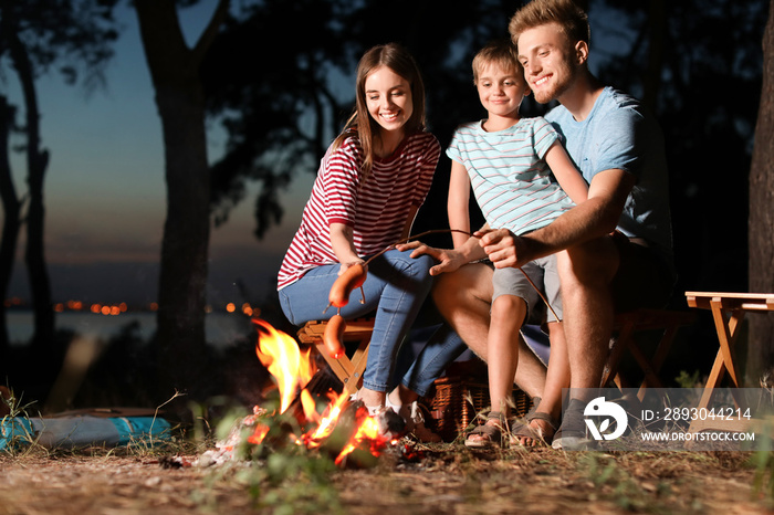 Family roasting sausages over campfire in evening