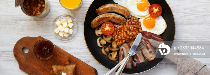 English breakfast in a frying pan with fried eggs, bacon, sausages, beans and toasts on white wooden