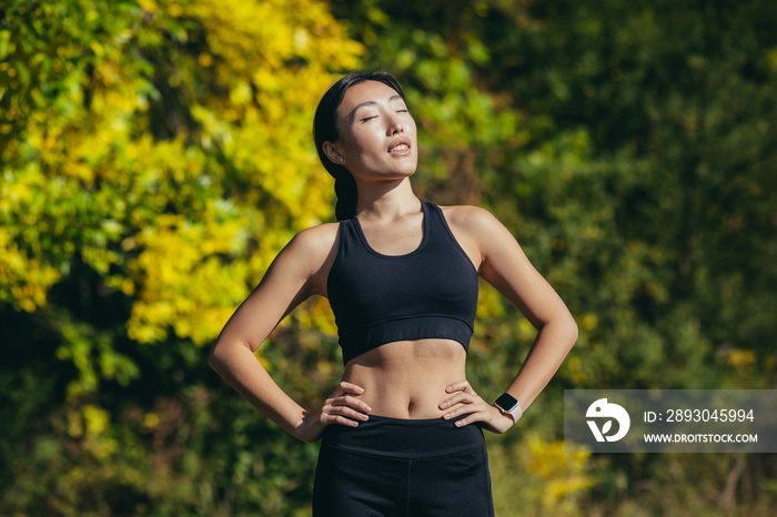 Close up portrait young happy fit asian woman standing in nature between forest trees relaxes, breat