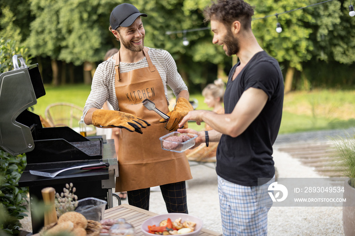 Two handsome male friends have fun while grilling meat for burgers on gas grill at backyard on natur