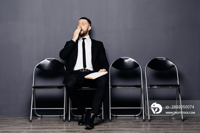 Serious bearded young man in glasses and formal wear is yawing while sitting in the hall on chair