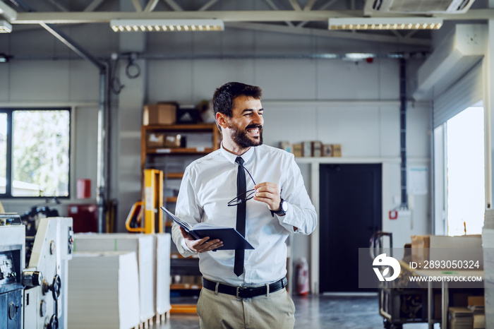 Cheerful caucasian bearded supervisor in shirt and tie holding notebook and eyeglasses while walking