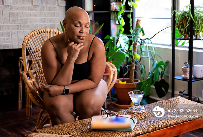 Bald Black woman sitting in wicker chair in living room surrounded by books and plants