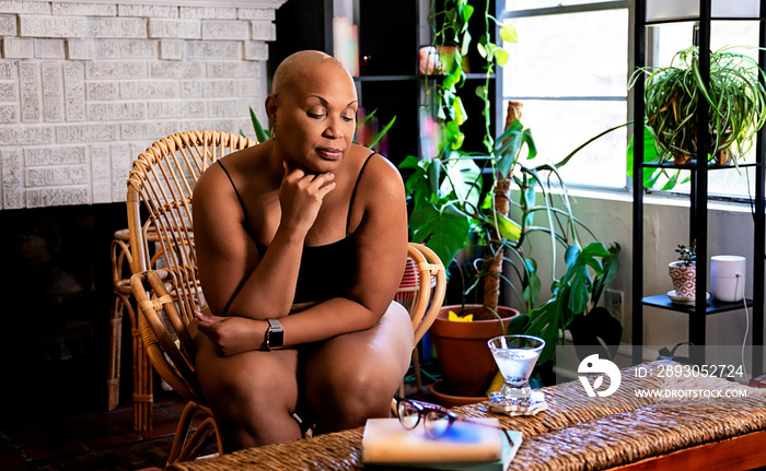 Bald Black woman sitting in wicker chair in living room surrounded by books and plants