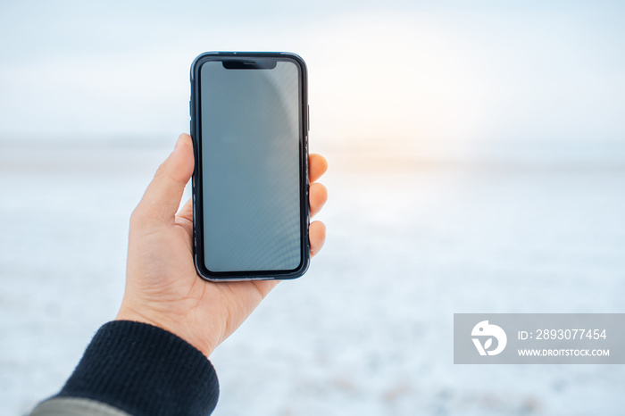 Close-up of male hand holding smartphone with mockup on background of blurred snowy field. Sunlight 