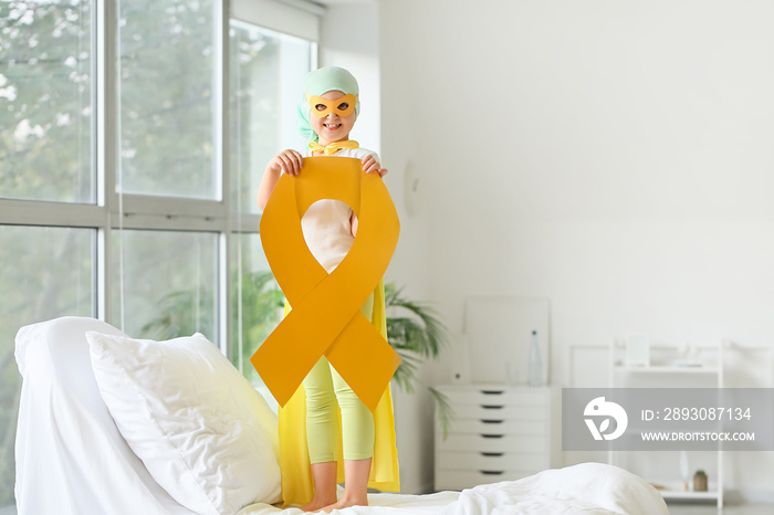 Little girl with golden ribbon wearing superhero costume in clinic. Childhood cancer awareness conce