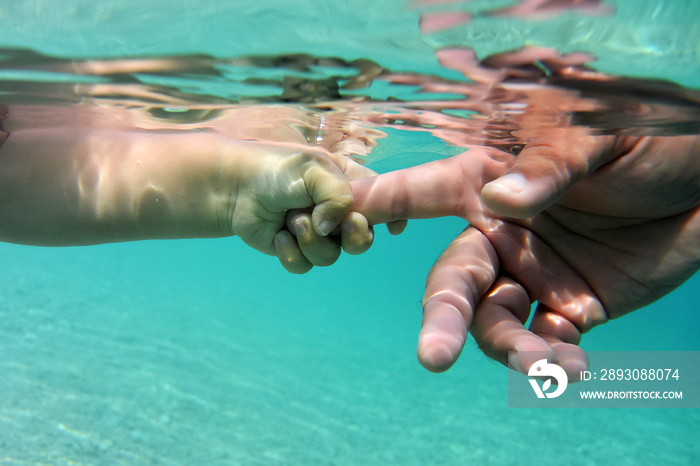 Baby hand holding fathers finger in transparent blue water. Underwater hands of father and small chi