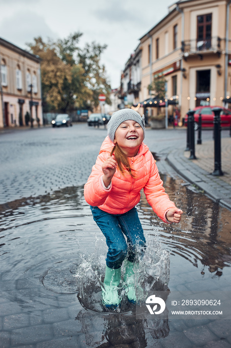 快乐的微笑女孩，在阴雨绵绵的秋日，在市中心散步时跳进水坑