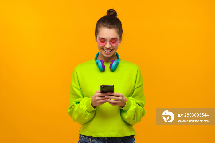 Young female wearing sweatshirt, colored eyeglasses and headphones around neck, looking at screen of