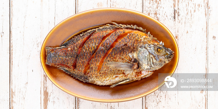 tasty large fried nile tilapia fish in oval ceramic plate on white wooden texture background with co