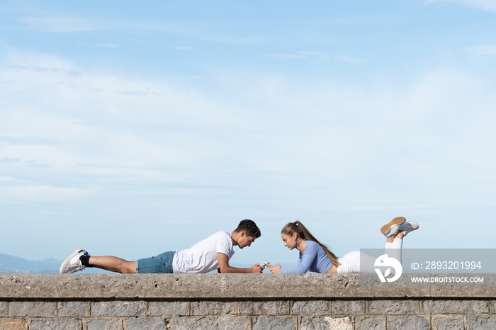 Teenager couple lying on a stone wall and using their phones. Digital addiction and dopamine concept