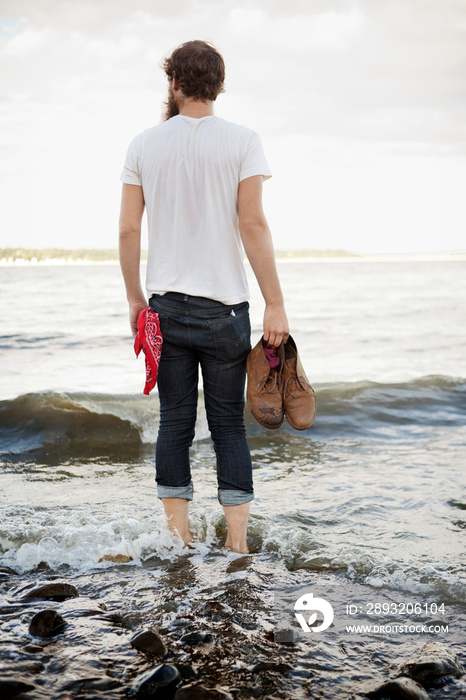 Rear view of man holding shoes while standing in water at beach