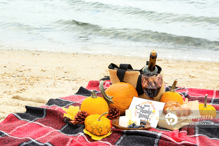 Pumpkins, delicious food, glass of wine and card with text HAPPY THANKSGIVING on beach near water