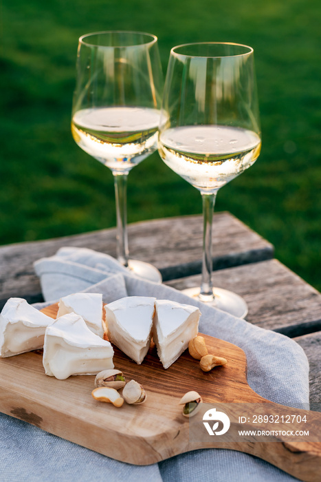 Two glasses of white wine and a wooden plate with cheese and nuts served outside at sunset.