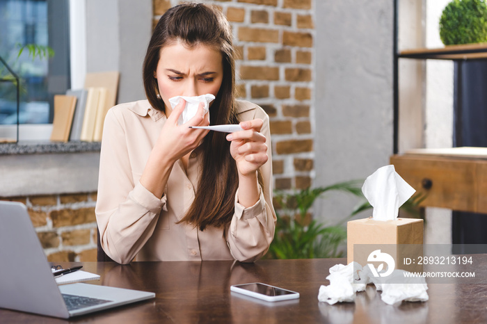 sick young businesswoman blowing nose and holding thermometer in office