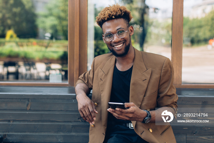 A portrait of African American man in jacket and glasses with smartphone sitting near the windows.