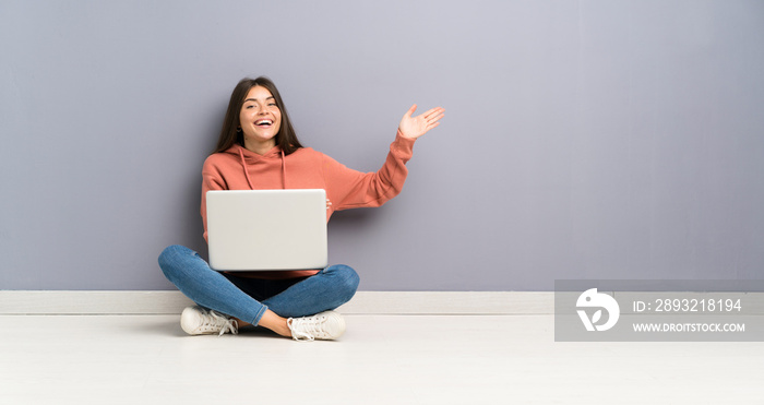 年轻的学生女孩在地板上拿着一台笔记本电脑，把手伸到一边，邀请她来