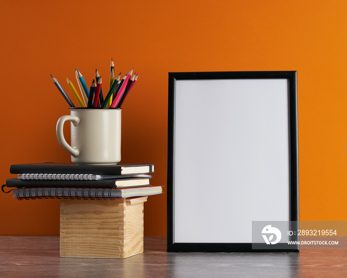 Desk with blank white picture frame and pencils in iron mug placed on books and wooden box. Orange b