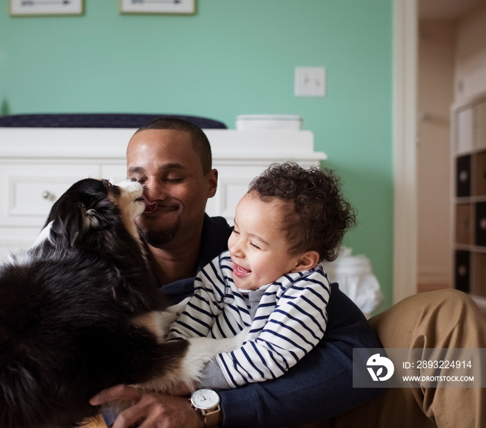 Playful dog licking father holding son while sitting against cabinet at home