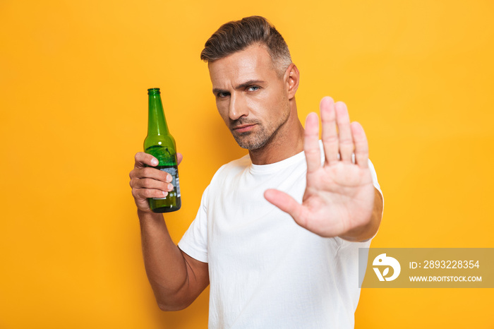 Image of displeased man 30s in white t-shirt holding bottle and drinking beer while standing isolate