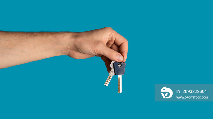 Unrecognizable young guy showing keys to new home on blue background, closeup. Blank space. Panorama